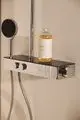 Duschsystem mit Brausethermostat in Chrom, montiert an einer Wand. Eine Duschhandbrause sowie eine Flasche Shampoo auf dem Ablagetablar. Ideal Standard ist die Marke.